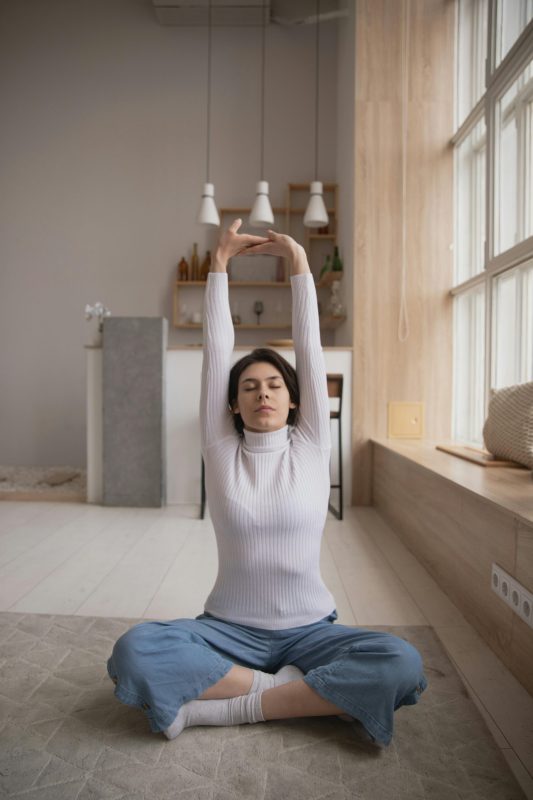Woman seated in yoga pose stretching arms upwards in a calm indoor setting.